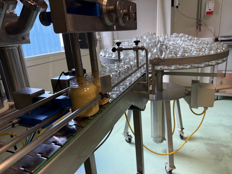 Vulmachine bij Mosterdmolen De Huisman die lege potten vult met grof gemalen mosterd, met een draaitafel vol schone potten op de achtergrond