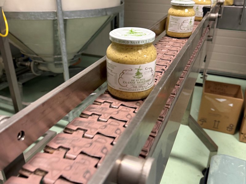 Gevulde potten ‘De Echte Zaanse Mosterd’ op een lopende band bij Mosterdmolen De Huisman, op weg naar de volgende verpakkingsstap